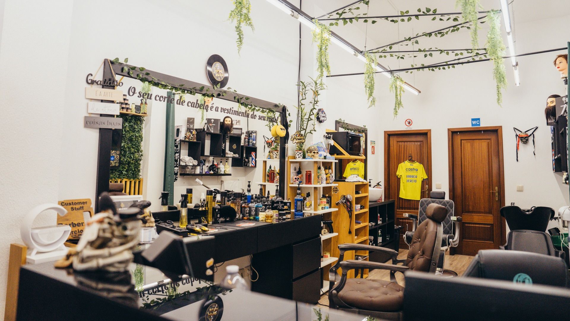 O interior da barbearia: espelho largo, prateleiras com produtos, plantas penduradas no teto e as cadeiras de corte.