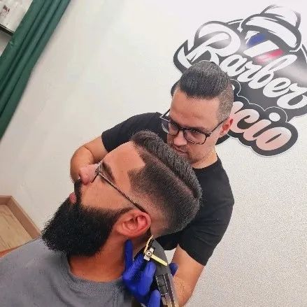 Cliente de barba cheia com contornos marcados e degradê nas laterais, enquanto o barbeiro acaba o corte.